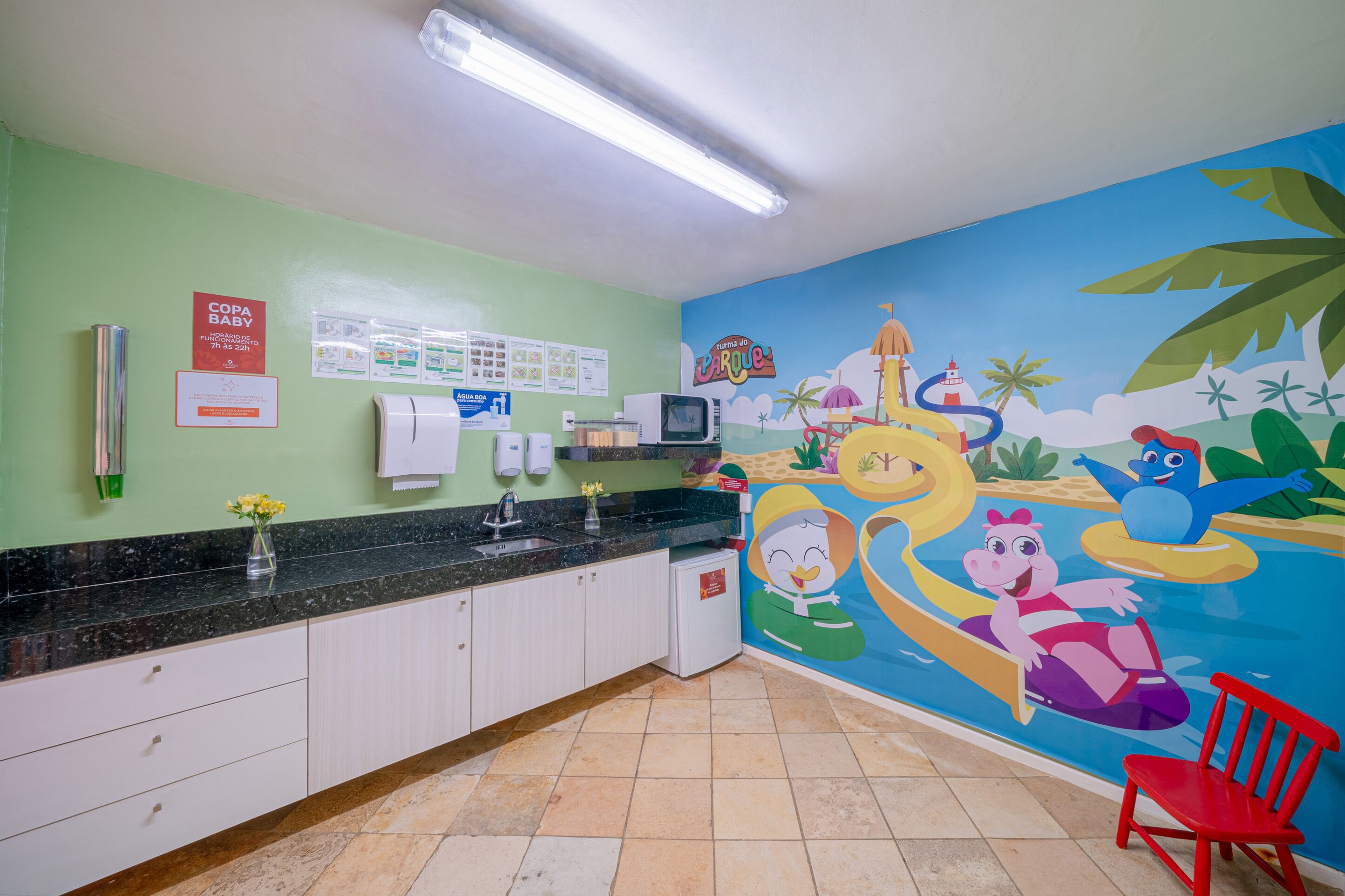 Spielbereich für Kinder mit Wandgemälde, Mikrowelle, Kühlschrank und einem roten Stuhl.