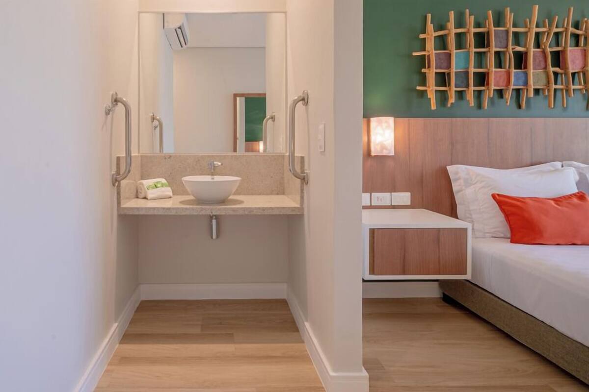 A bathroom with a sink, mirror, and handrails. The room has a bed with white bedding and a red pillow.