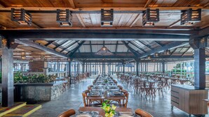 A spacious indoor dining area with long tables set for multiple guests, wooden beams on the ceiling, and a view through large windows.