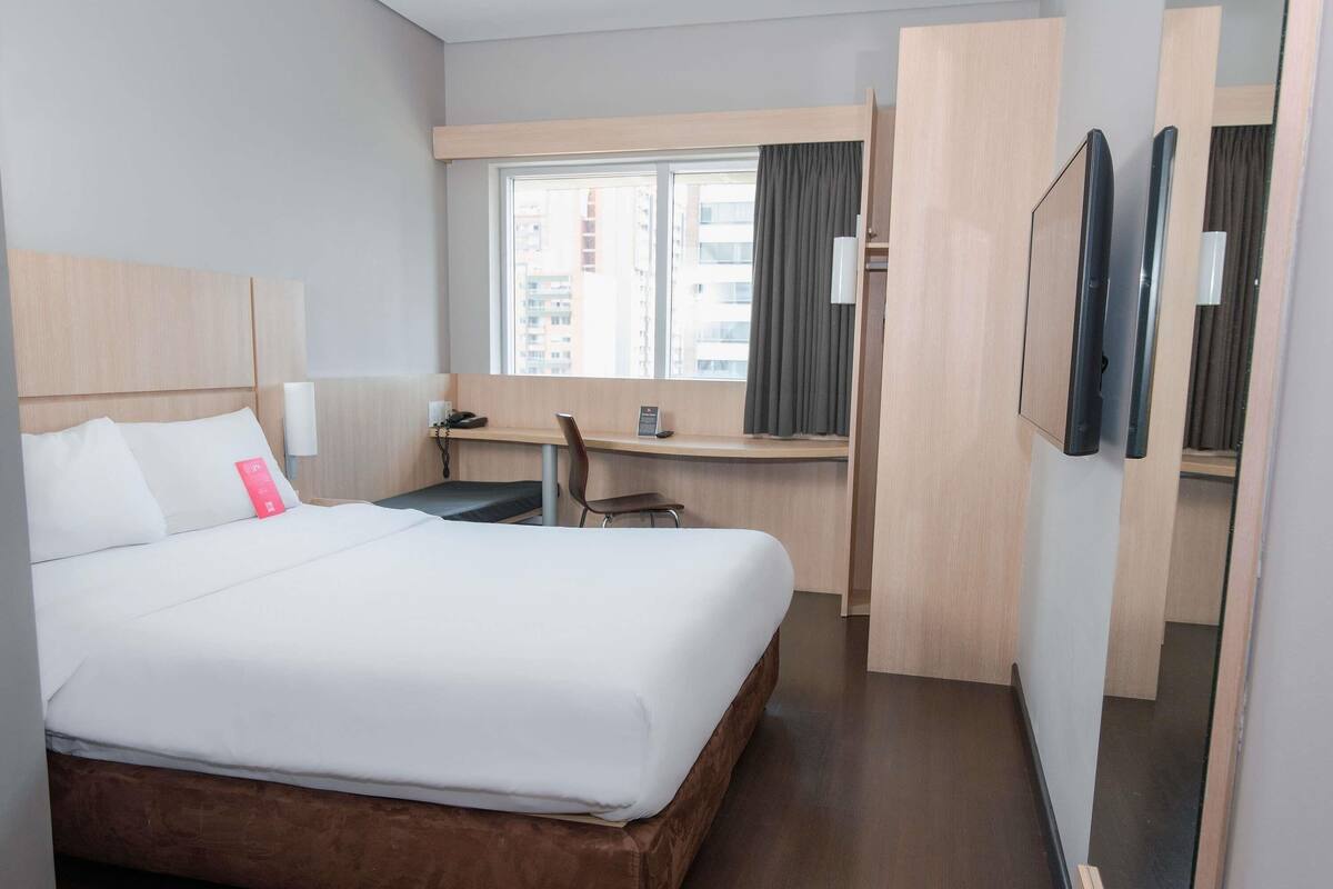 Quarto de hotel com uma cama grande, uma escrivaninha, uma cadeira, um telefone e uma janela com cortinas.