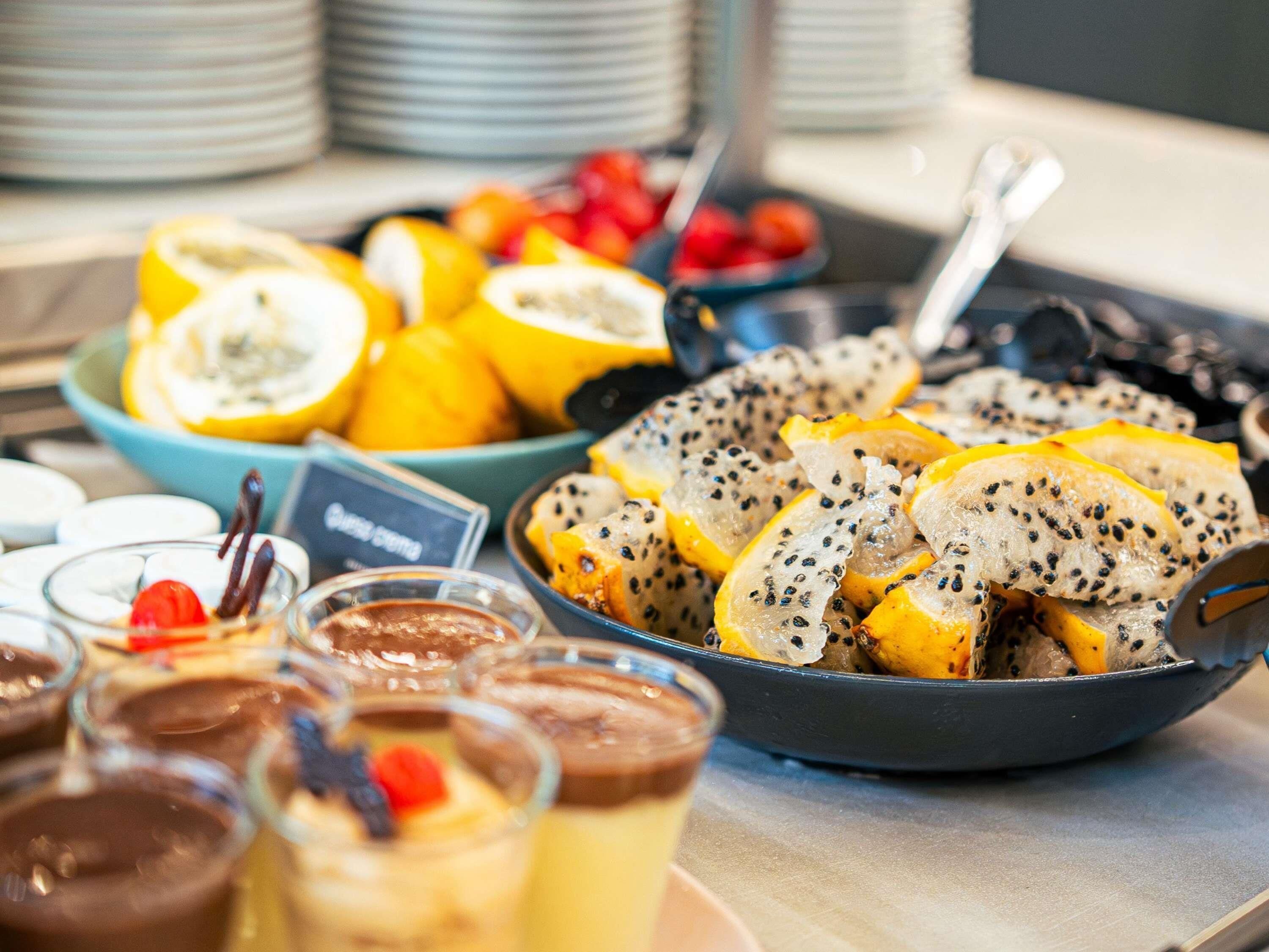 Un bufé con frutas en rodajas, una bandeja de pan con semillas y diversos postres.