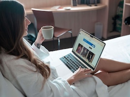 A person using a laptop in a hotel room with a bed, a chair, and a small table.