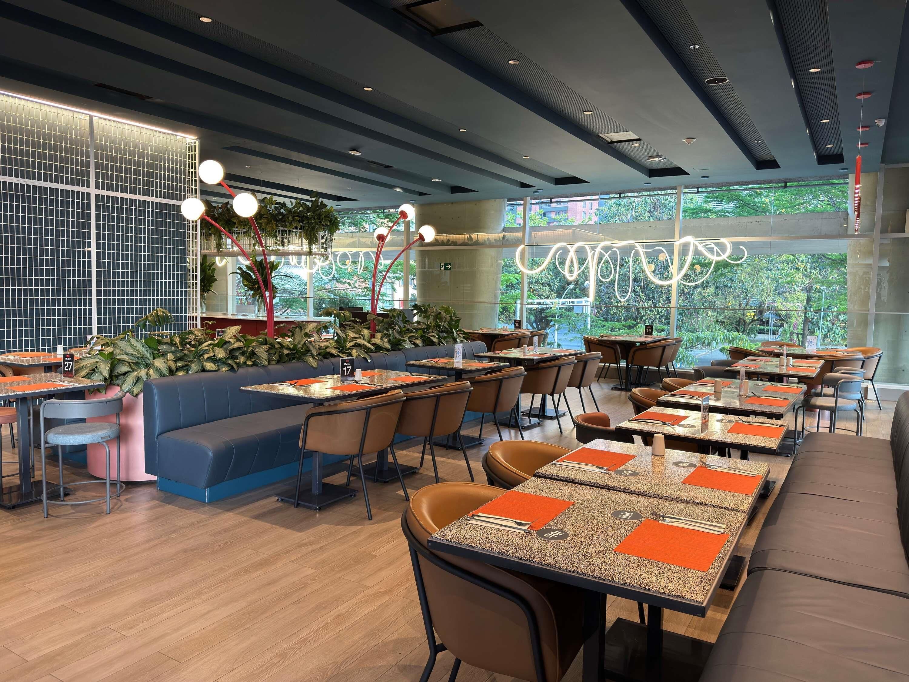 Ein modernes Restaurant mit hohen Tischen, Sitzecken und einem großen Fenster mit Blick auf Grünflächen.