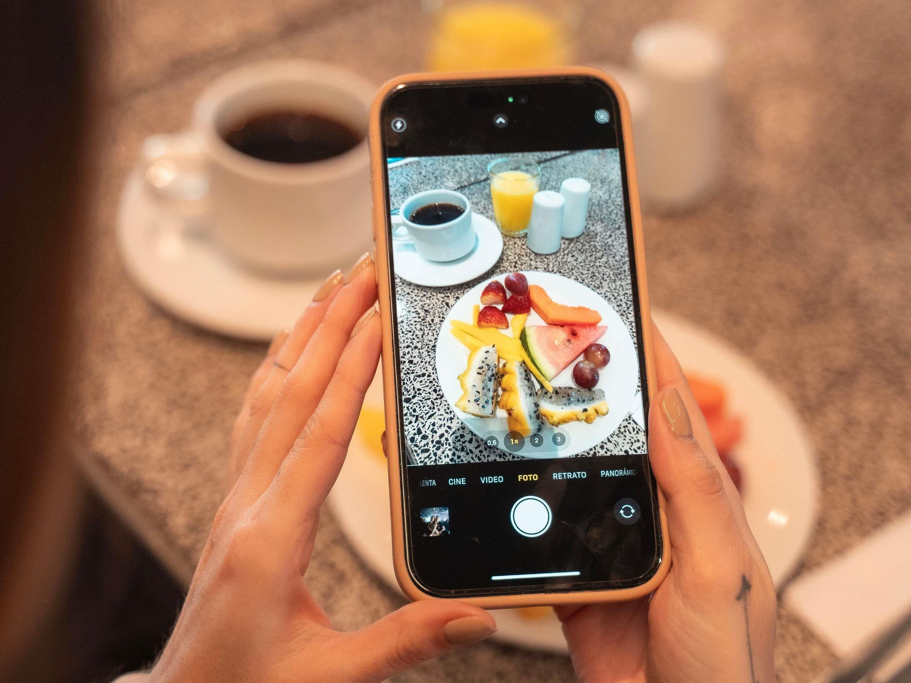 Une personne tient un smartphone sur lequel est affichée une photo d’une assiette de petit-déjeuner contenant divers fruits et une tasse de café.