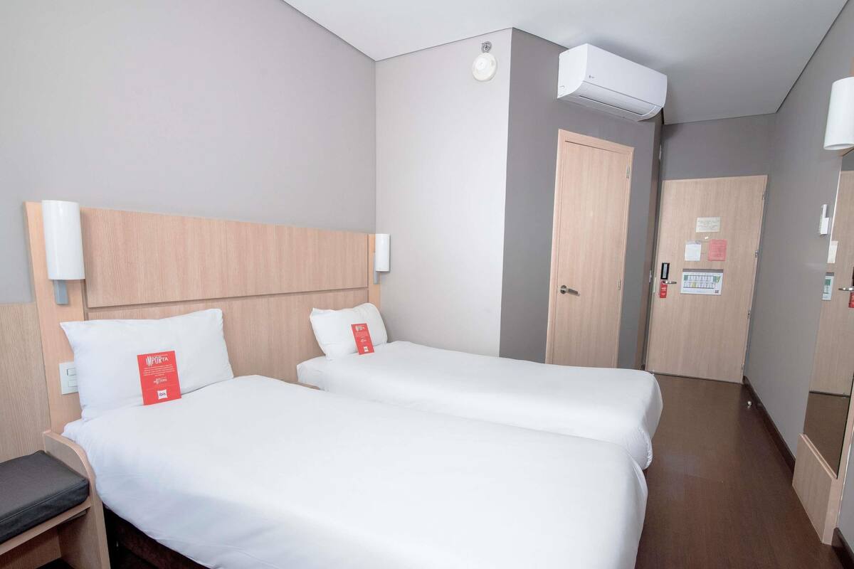 Quarto de hotel com duas camas, cabeceira de madeira, mesa de cabeceira, uma cadeira, guarda-roupa e ar condicionado.