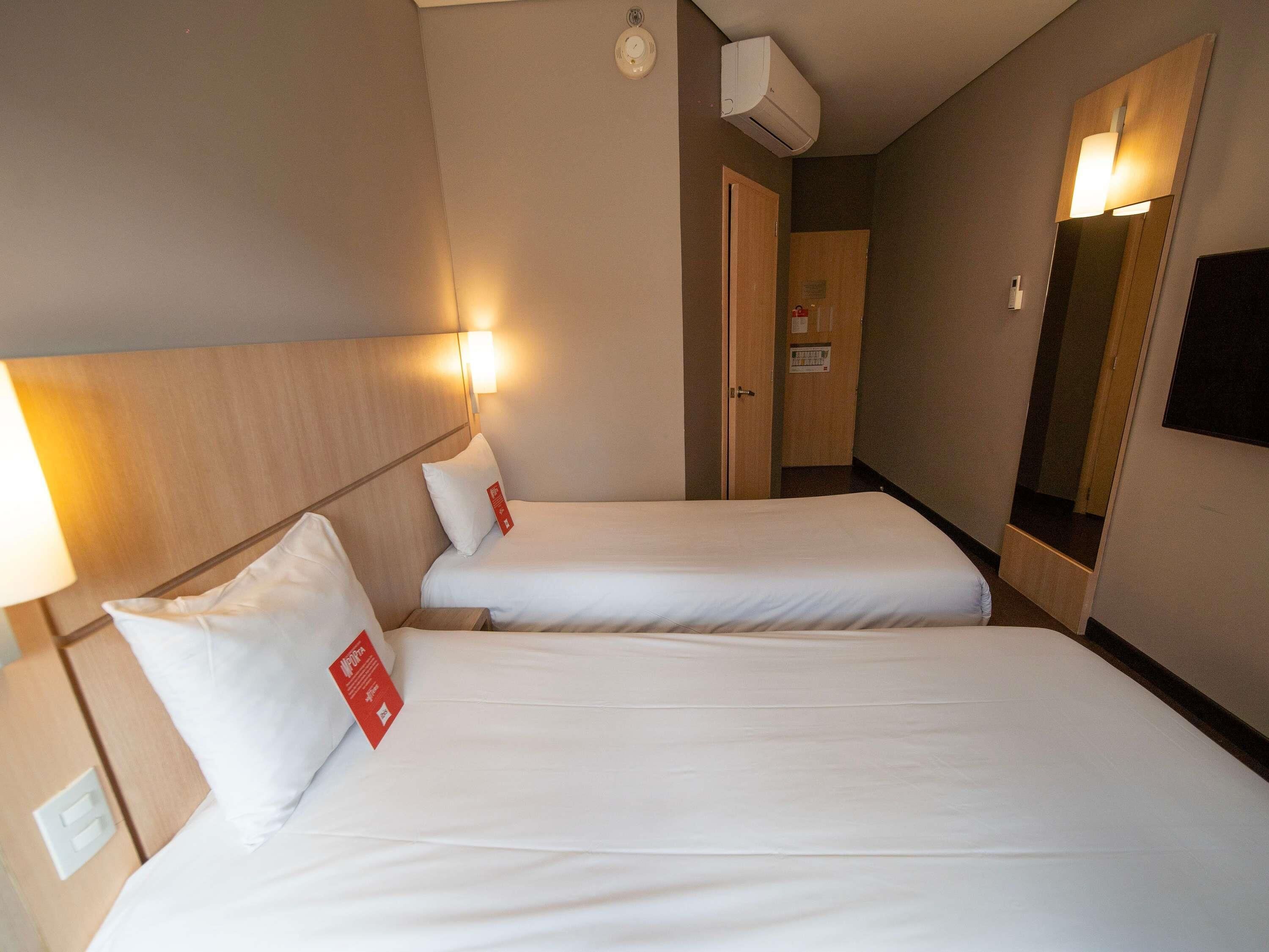 A hotel room with two single beds, a bedside table with a lamp, a wall-mounted air conditioner, and a mirror.