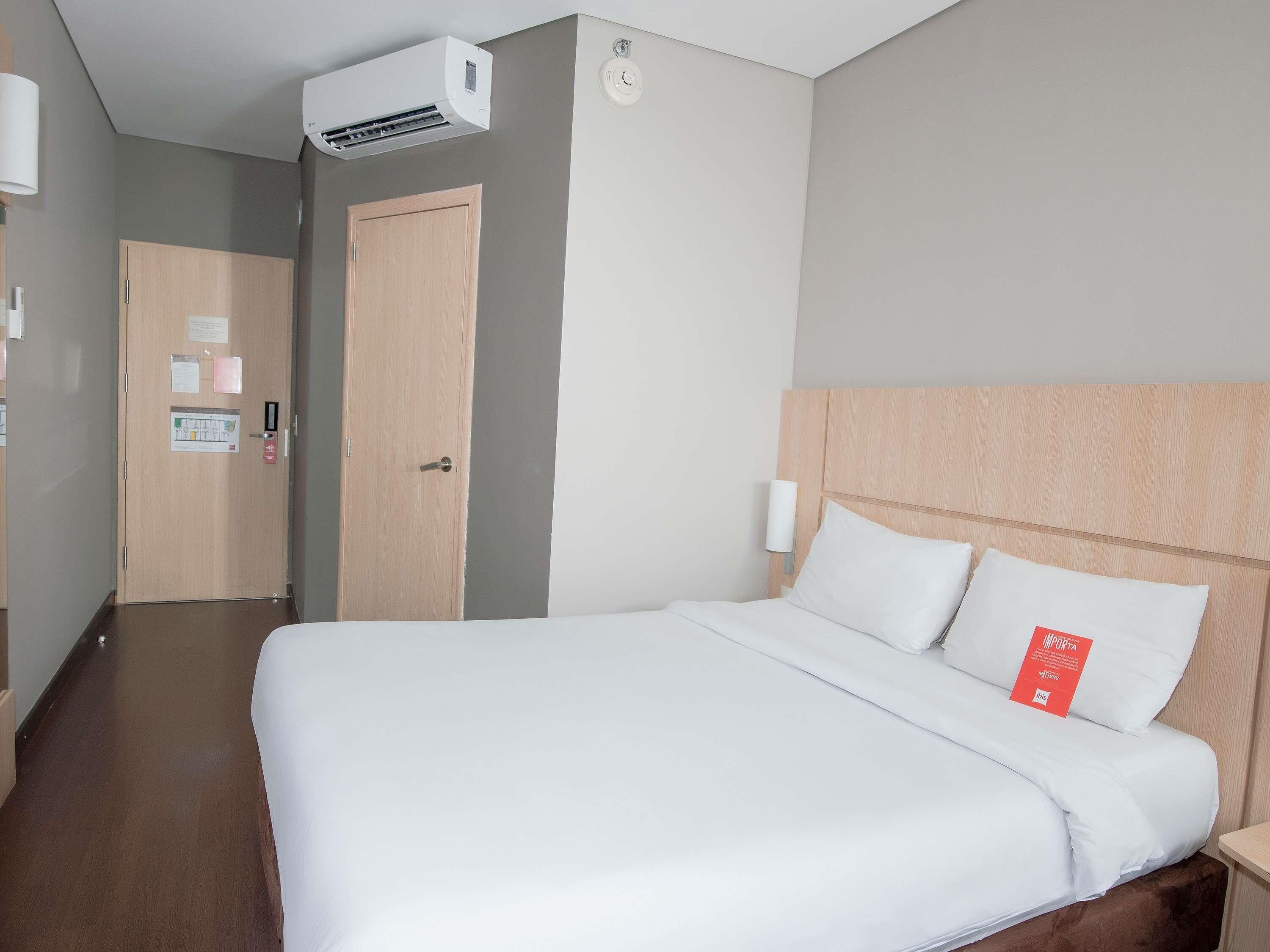 Quarto de hotel com cama, cabeceira de madeira, ar-condicionado fixado na parede e porta com uma placa.