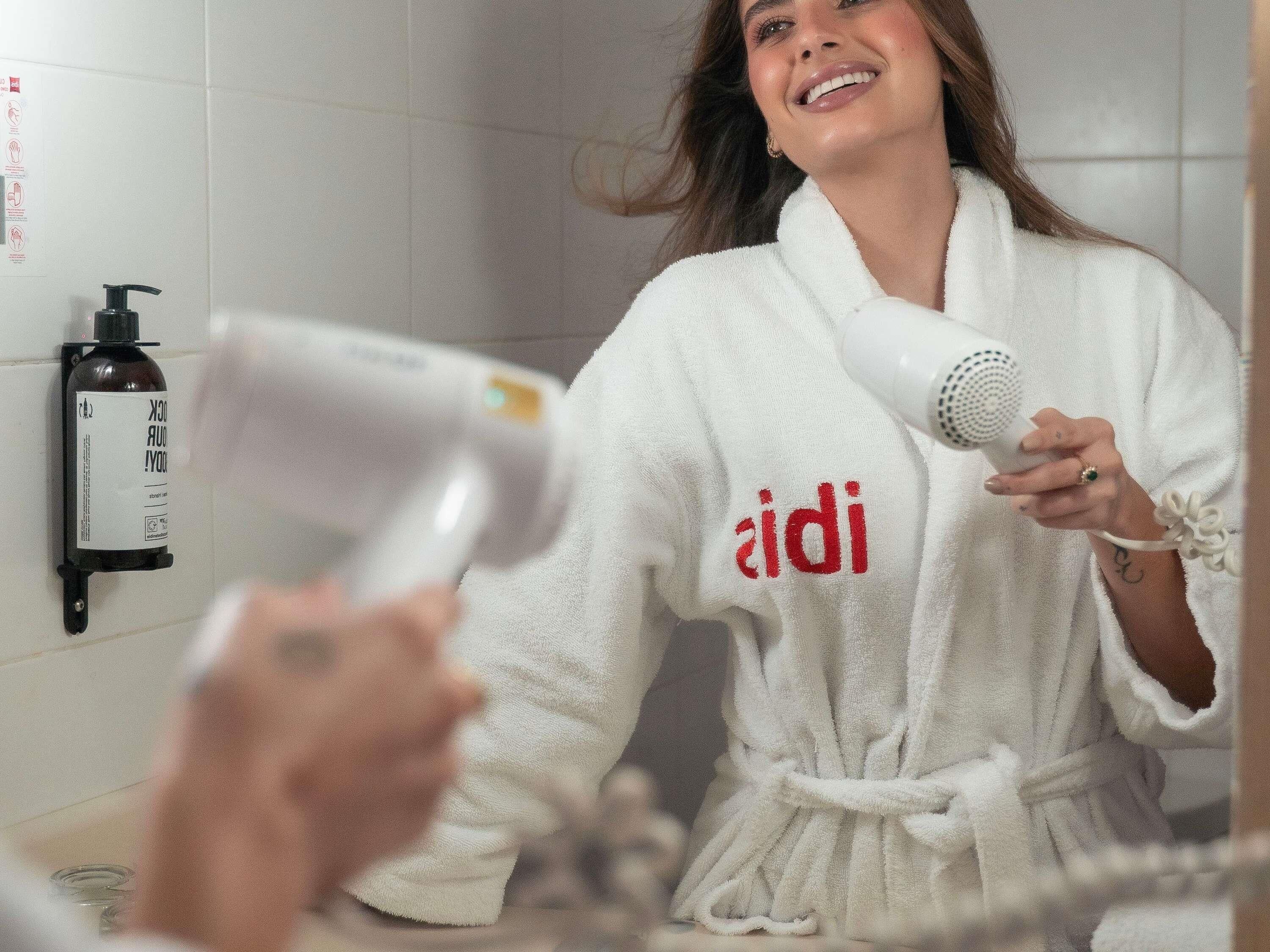 Una persona vestida con una bata blanca con el texto 'aidi', sosteniendo un secador de pelo en un baño con pared blanca y una botella de desinfectante de manos.