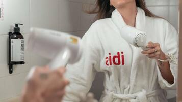 Una persona vestida con una bata blanca con el texto 'aidi', sosteniendo un secador de pelo en un baño con pared blanca y una botella de desinfectante de manos.