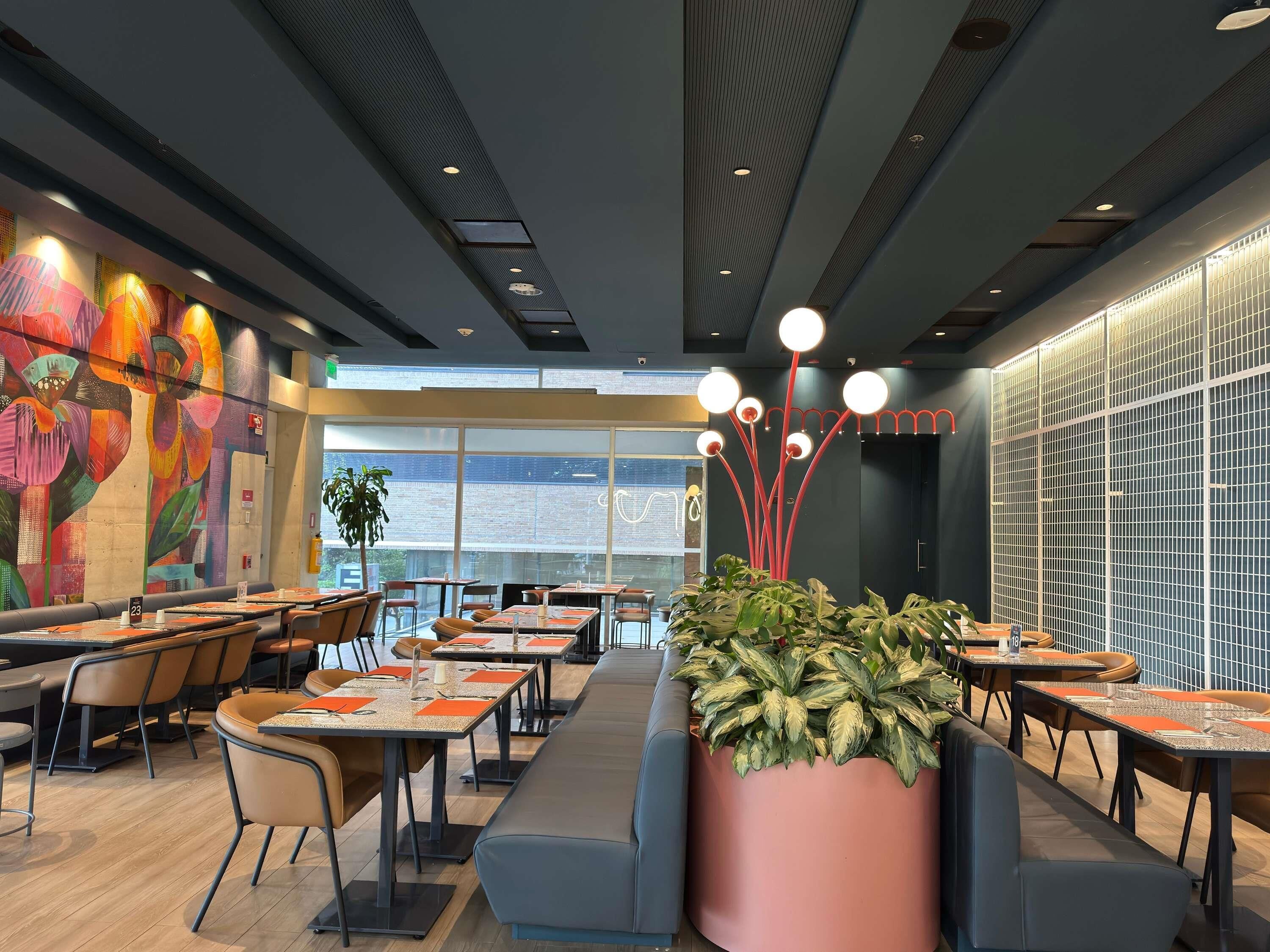 Een modern restaurantinterieur met hoge plafonds, grote ramen en een kleurrijke muurschildering aan de linker muur.