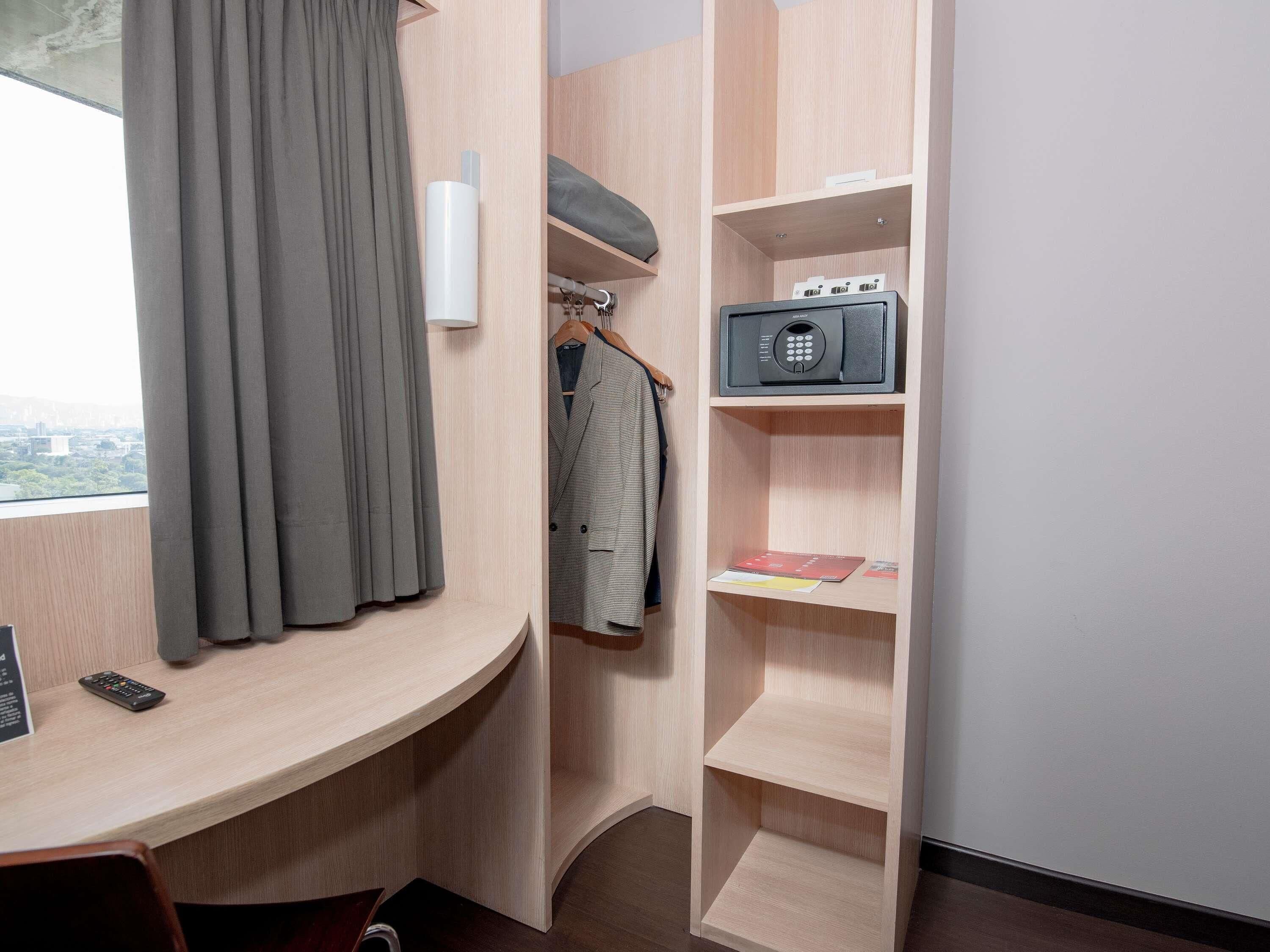 Habitación de hotel con un escritorio curvo, perchero con una chaqueta, una caja de seguridad pequeña y una ventana con cortinas.