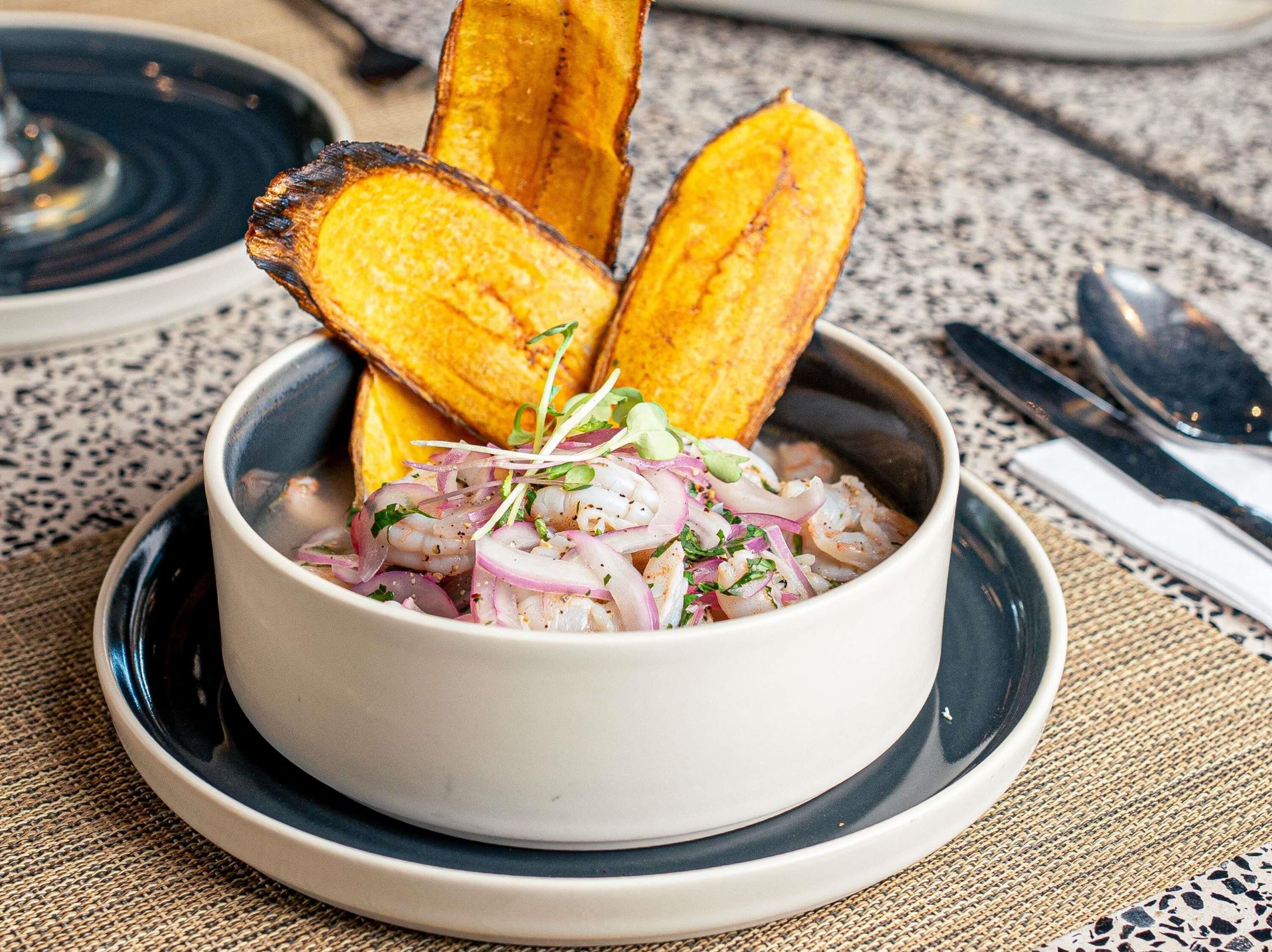 A bowl of shrimp cocktail with red onions and herbs, accompanied by grilled plantain slices on a plate.