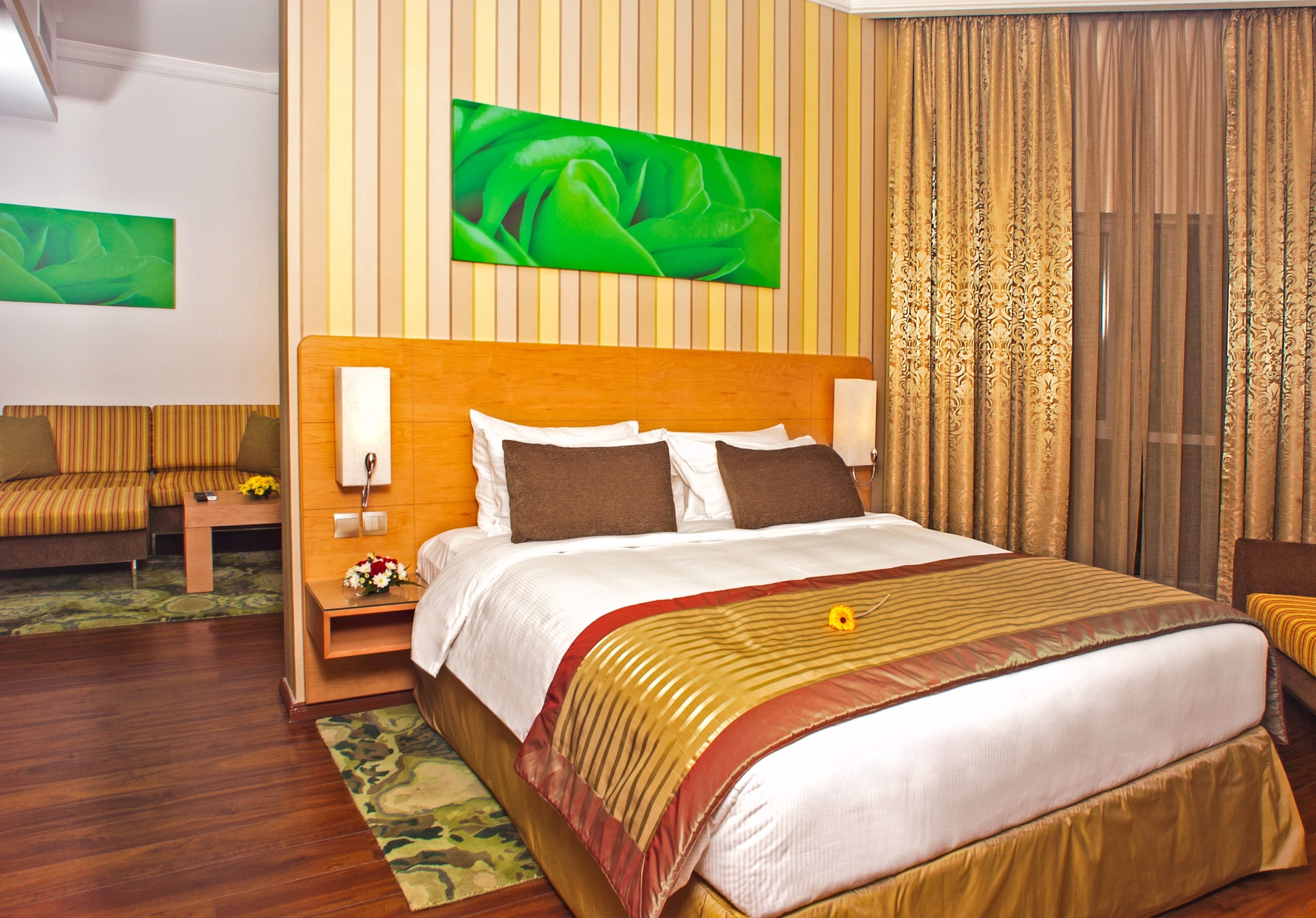 A hotel room with a large bed, a wooden headboard, a nightstand with a lamp, a sofa, and a painting on the wall.