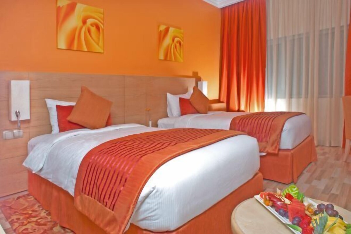 A hotel room with two beds, orange bedding, a fruit platter on a side table, and artwork on the walls.