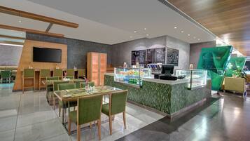 A modern restaurant with a central counter, green upholstered chairs, and a menu board.