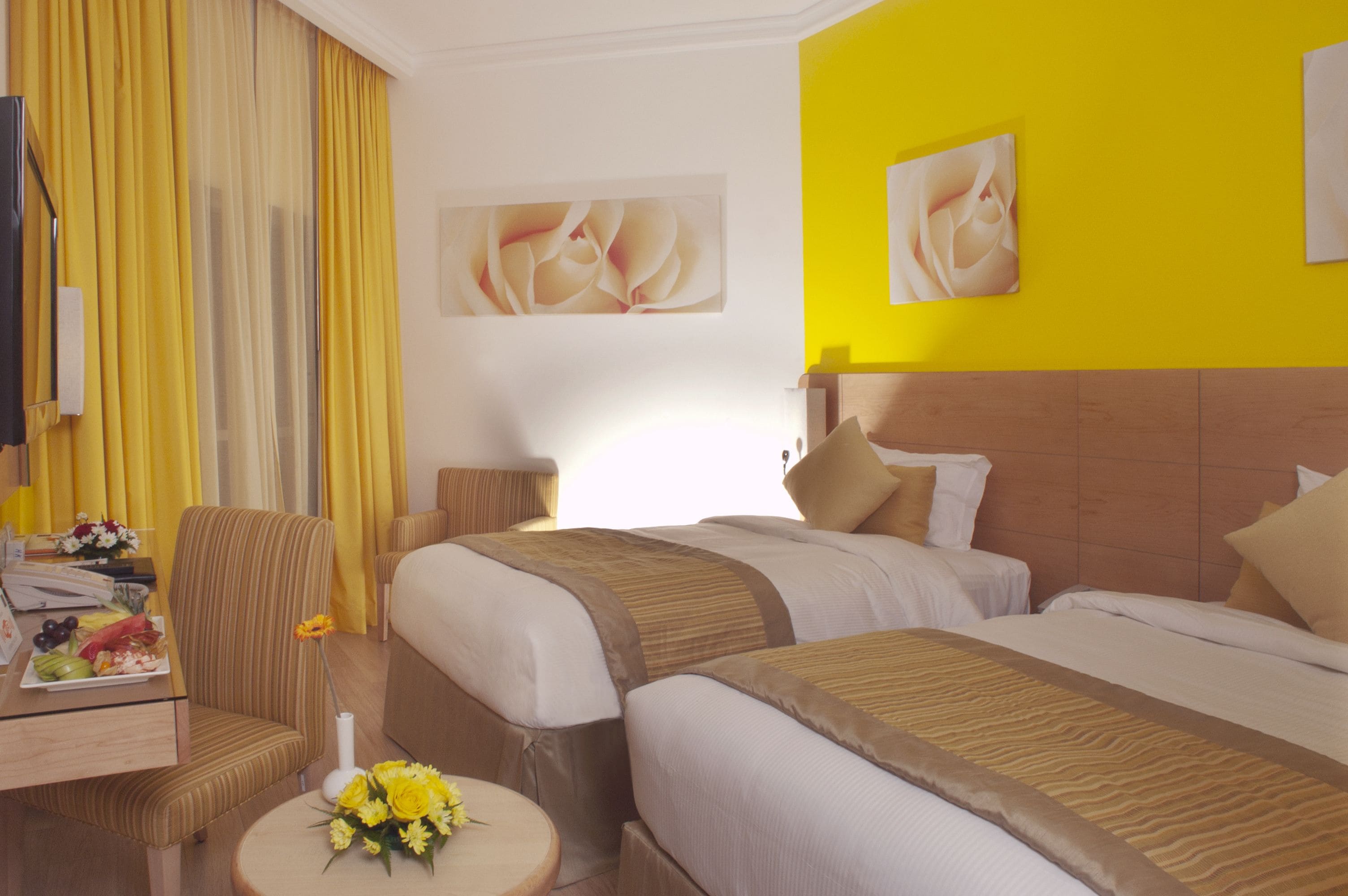 Habitación de hotel con dos camas, un escritorio con silla, una mesita con flores y obras de arte en las paredes.