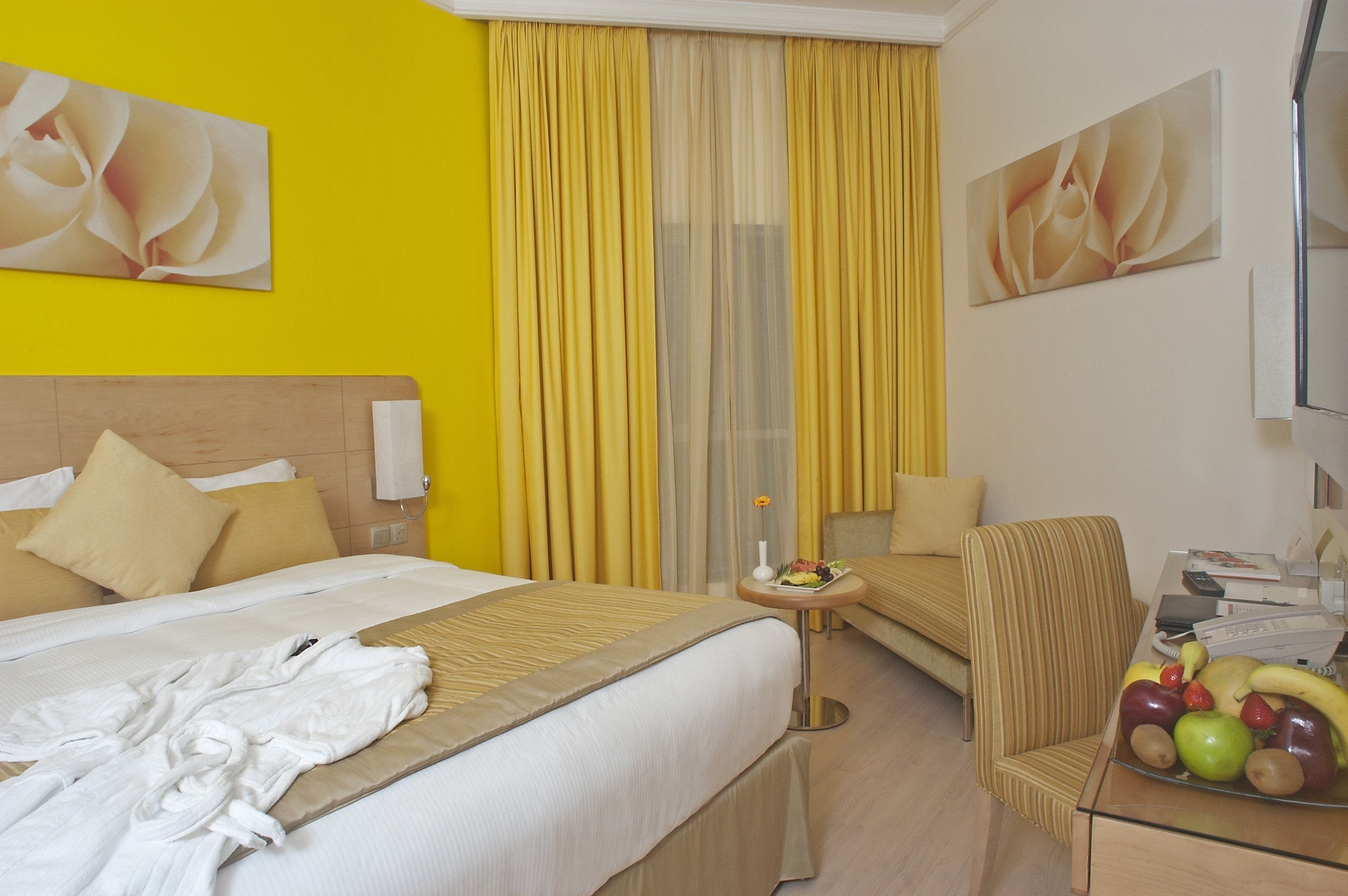 A hotel room with a bed, a bedside table with a fruit bowl, a chair, and a small table with a vase.