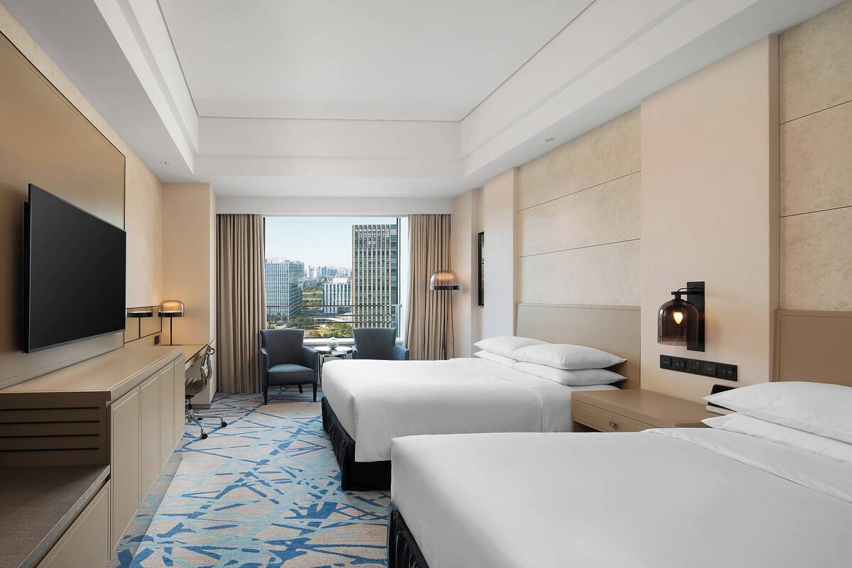 A hotel room with two beds, a desk, a TV, a chair, and a view of the cityscape.