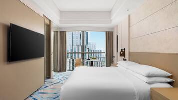 Una habitación de hotel con una cama grande, un televisor de pantalla plana, una mesita con una planta y vistas a edificios altos a través de amplios ventanales.