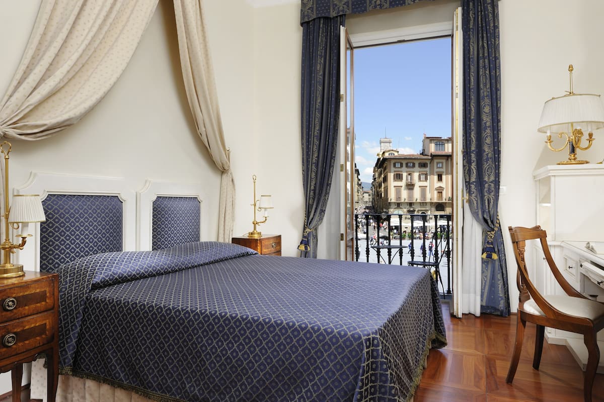 A bedroom with a bed, a wooden nightstand, a chair, and a view of buildings through a balcony door.