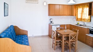 A compact kitchen with wooden cabinets, a dining table with chairs, a coffee machine, and a small refrigerator. - Fotini Apartments (Sitia)