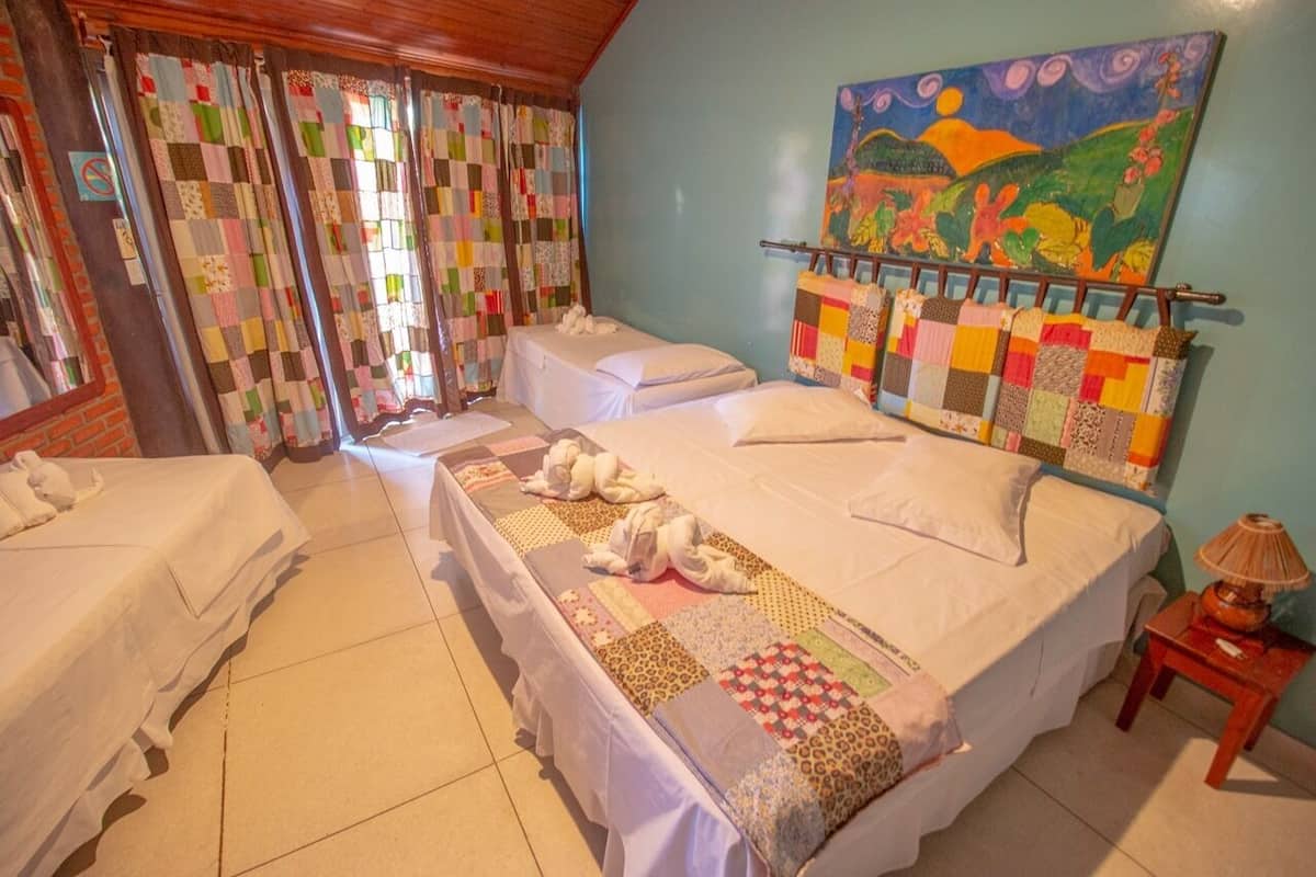 A hotel room with two beds, a quilted curtain, a painting, and a lamp.