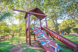 Un parque infantil con tobogán y columpios, rodeado de árboles y césped.