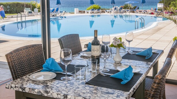 Une table dressée pour un dîner à deux, avec une bouteille de vin, des verres et un vase de fleurs, donnant sur une piscine et des chaises longues.
