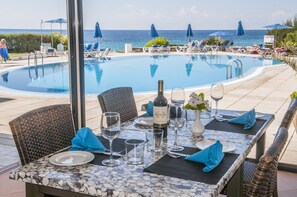Une table dressée pour un dîner à deux, avec une bouteille de vin, des verres et un vase de fleurs, donnant sur une piscine et des chaises longues.