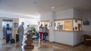 Un vestíbulo de hotel con una recepción, mostrador de registro, zona de estar con una mesa y sillas, y un gran ventanal que permite la entrada de luz natural.