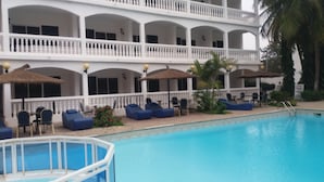 A poolside area with lounge chairs and umbrellas, a pool, and a multi-story building with balconies. - Sunu Hotel (Serrekunda)