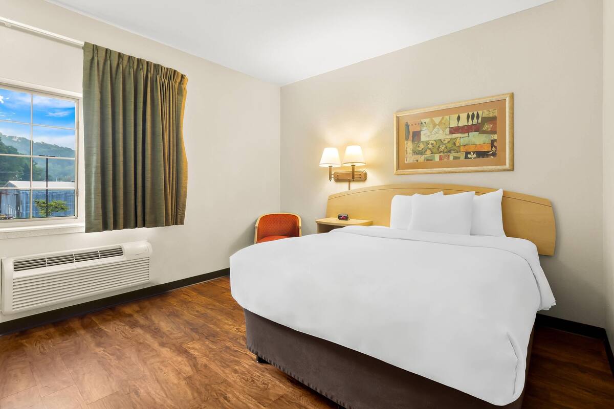 A hotel room with a large bed, a window with curtains, a wall-mounted lamp, and a painting.
