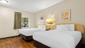 A hotel room with two beds, a window with curtains, a wall-mounted lamp, and a painting. - Suburban Studios Morgantown (Morgantown)