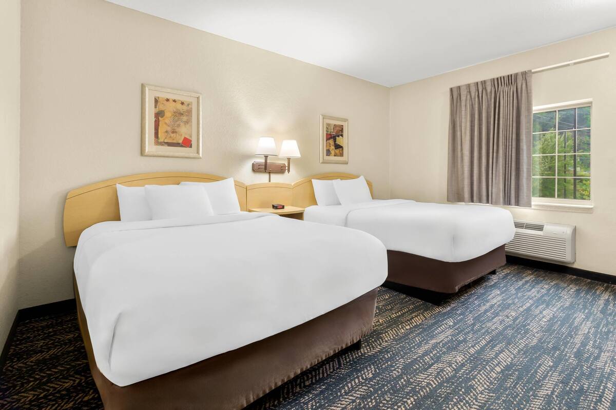 A hotel room with two beds, a large window with curtains, a wall-mounted lamp, and a framed picture.