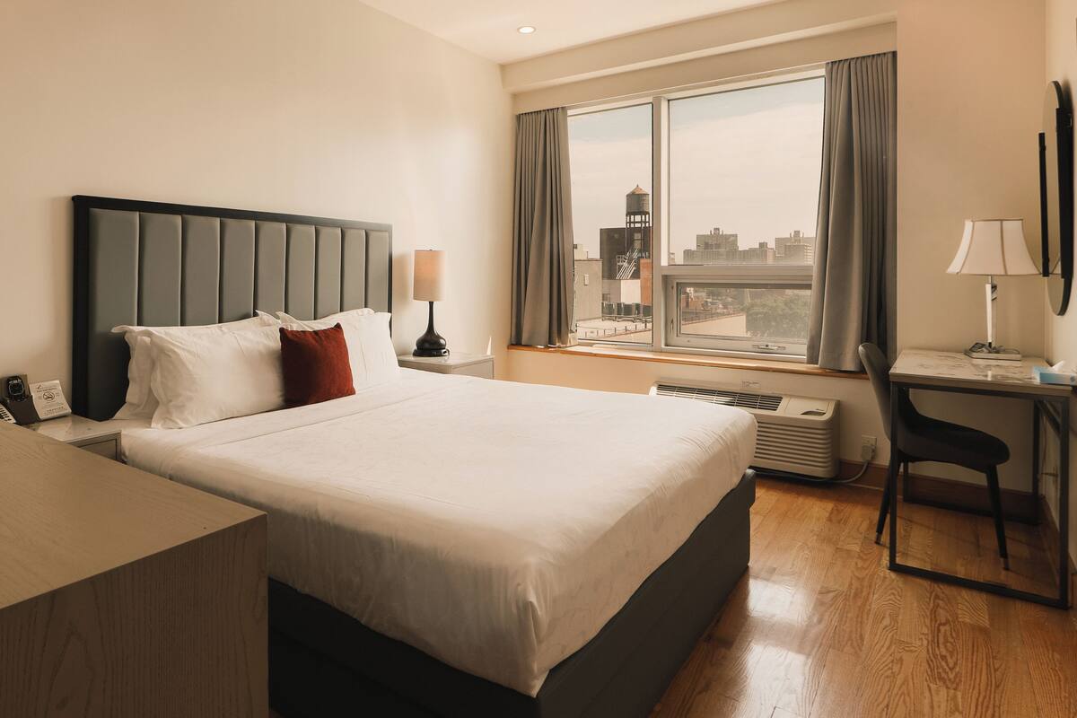 A hotel room with a large bed, a desk, a chair, and a window with a city view.