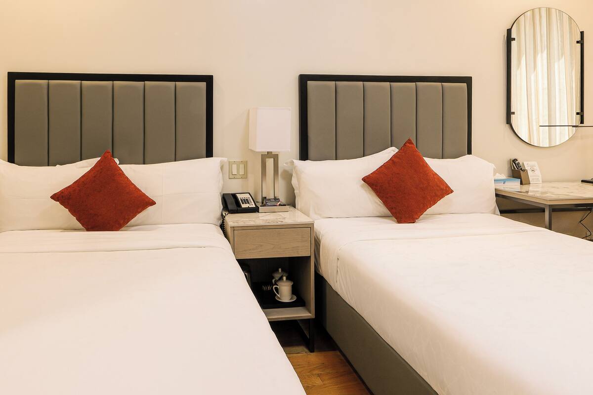A hotel room with two beds, each with a headboard, a nightstand with a lamp, a small table with a phone and a remote control, and a wall-mounted mirror.