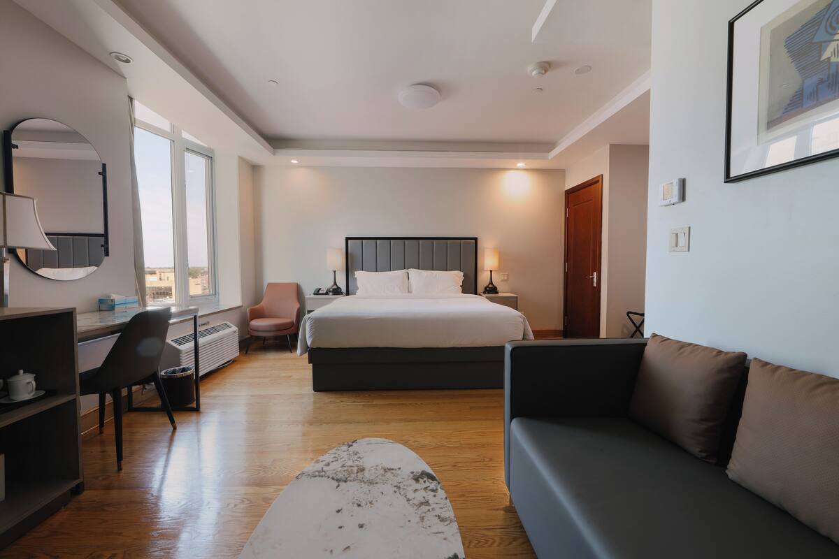 A modern hotel room with a large bed, a desk, a chair, a sofa, and a large window.