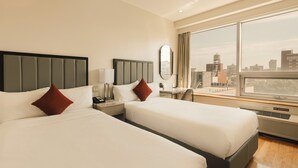 A hotel room with two beds, a desk, a chair, a window with a city view, and a 'No Smoking' sign.