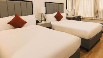 Quarto de hotel com duas camas, cada uma com lençóis brancos e almofada vermelha. Há uma mesa de cabeceira com luminária, telefone e espelho. O quarto possui piso de madeira e uma janela com cortinas.