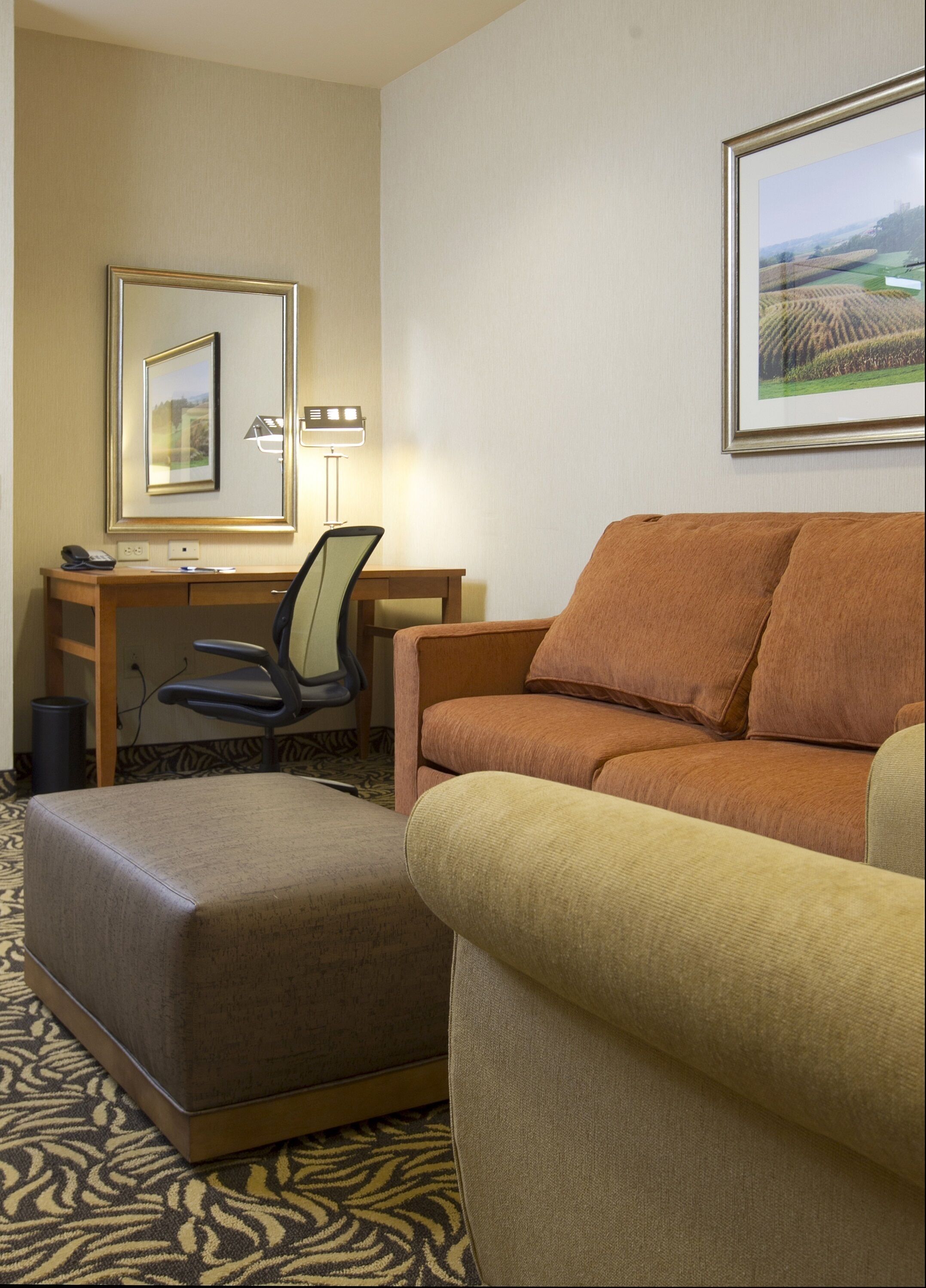 A hotel room with a brown sofa, a matching ottoman, a desk with a chair, a lamp, and a framed picture on the wall.