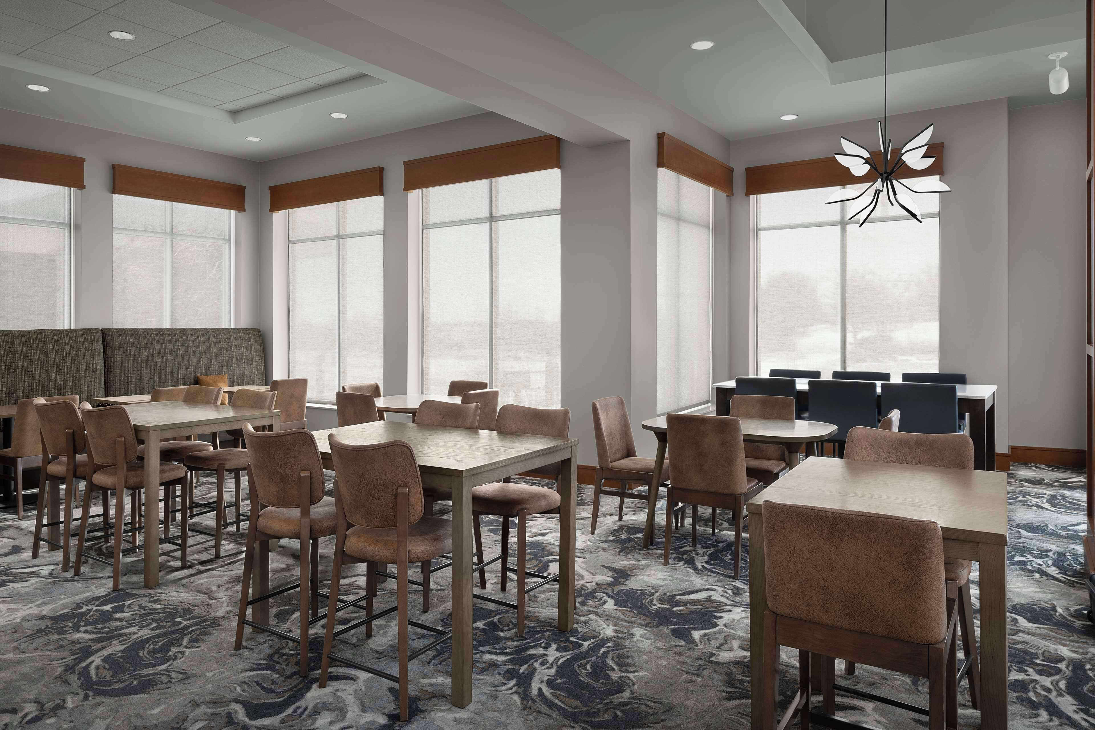 A hotel dining area with multiple tables and chairs, large windows, and a marble-patterned carpet.