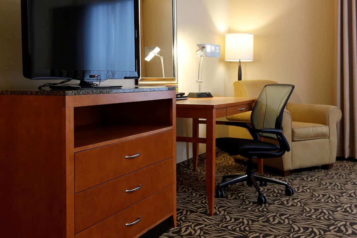 A hotel room with a wooden desk, a television, a mirror, a lamp, a chair, and a sofa.