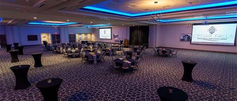Un salón de conferencias con un escenario, varias mesas y sillas dispuestas para un evento.