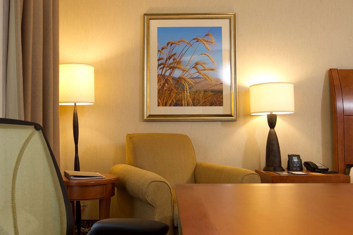 A hotel room with a single yellow armchair, a wooden side table with a lamp, a framed picture of wheat on the wall, and a bed with a white headboard.