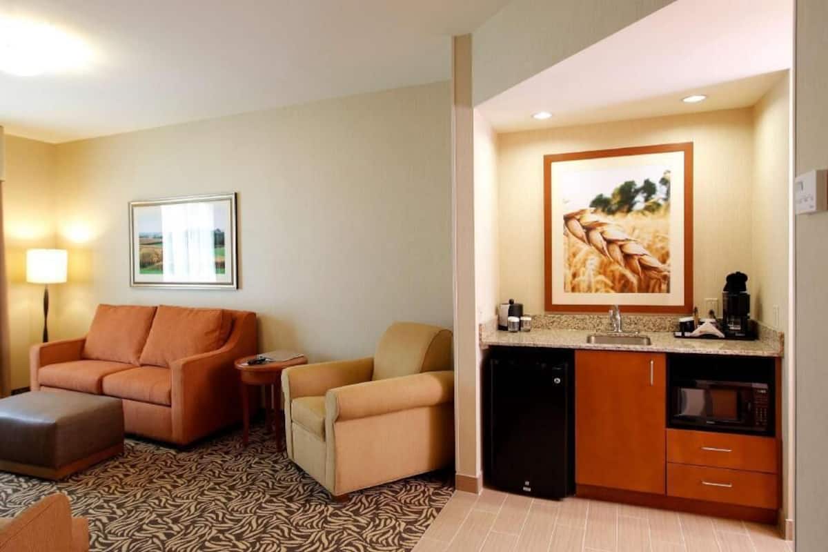 A hotel room with a sofa, armchair, ottoman, kitchenette, and a framed picture on the wall.