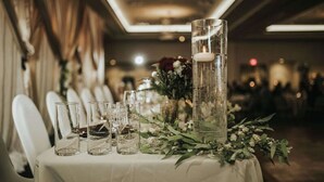 A table set with clear glasses and a tall glass vase with a candle, surrounded by greenery and flowers. - Hilton Garden Inn Cedar Falls (Cedar Falls)