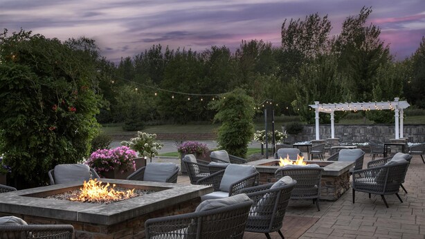 An outdoor patio with stone fire tables, wicker chairs, and a pergola with string lights. - Hilton Garden Inn Cedar Falls (Cedar Falls)