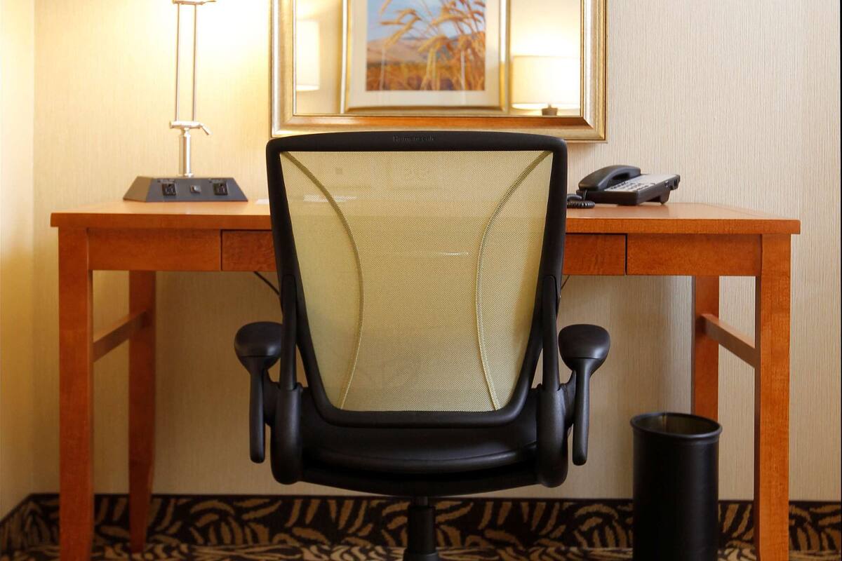 A hotel room with a wooden desk, a black office chair, a lamp, a framed picture, and a telephone.