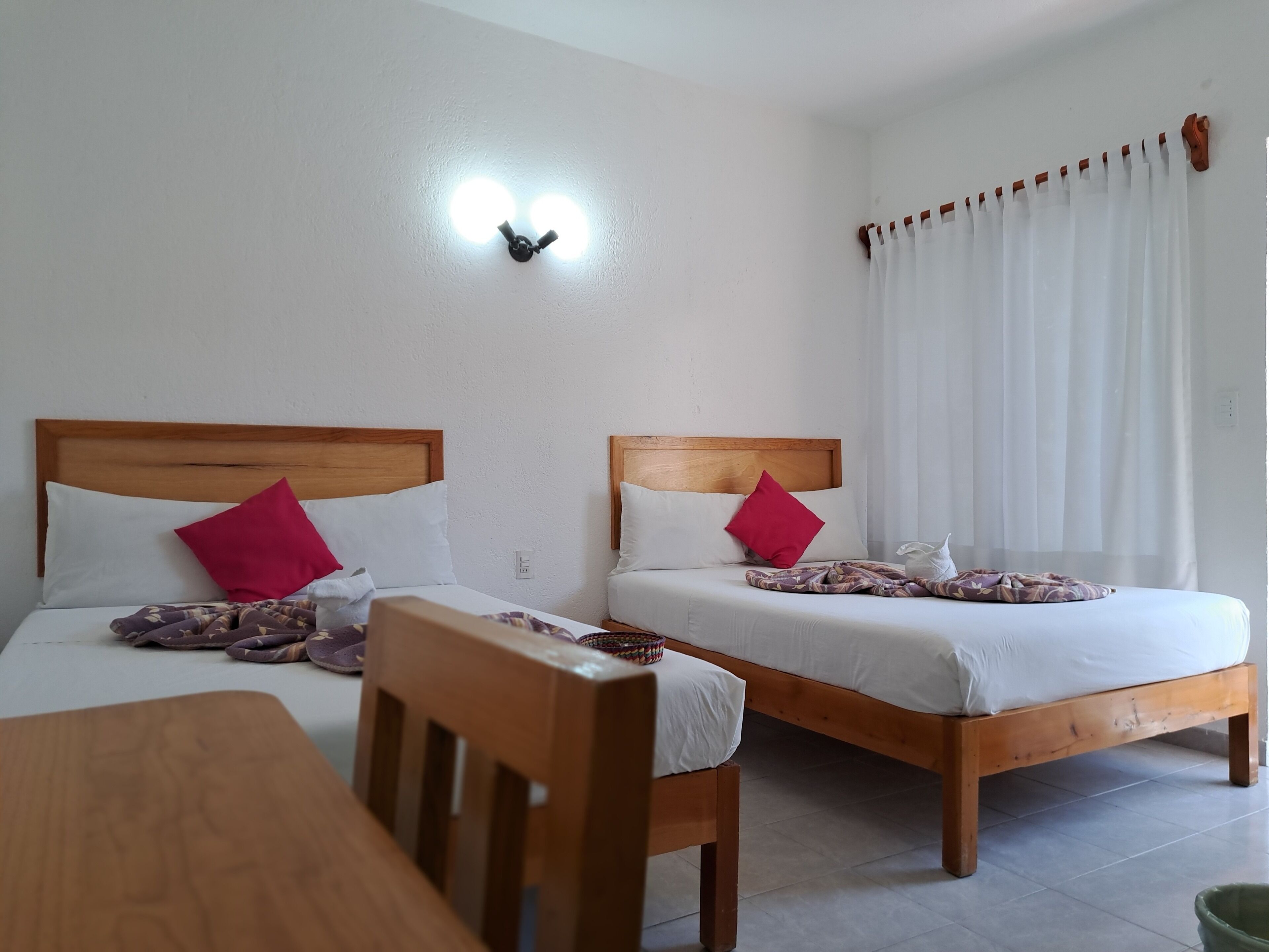 Una habitación de hotel con dos camas de madera, ropa de cama blanca, almohadas rojas, una silla de madera y una mesa.