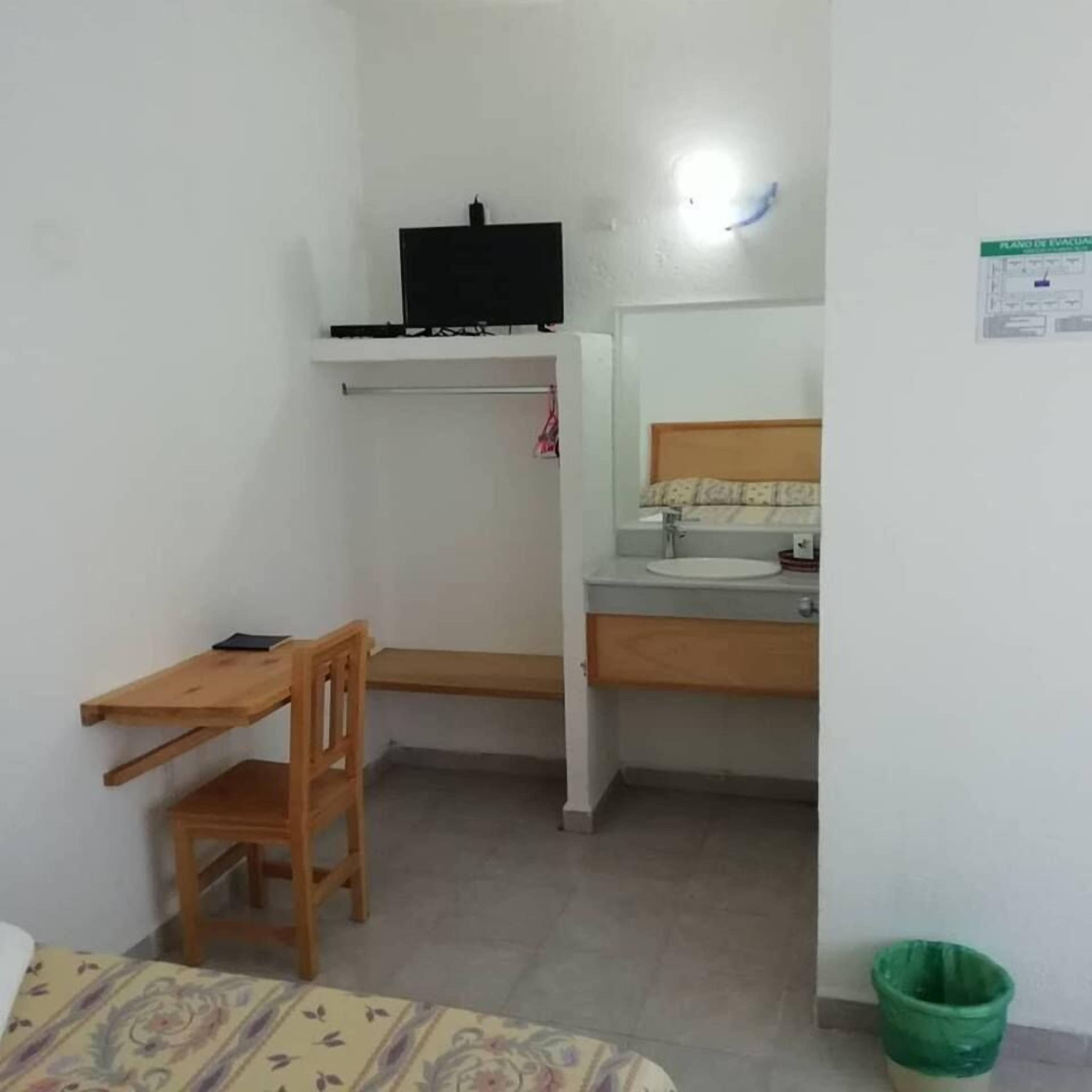A small room with a bed, a desk with a chair, a TV mounted on the wall, a sink, and a green trash bin.